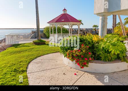 Belvédère en bois blanc avec toit rouge se trouve sur une pelouse verte luxuriante surplombant l'océan Pacifique à Oceanside, Californie, entouré d'un jardin tropical landsc Banque D'Images