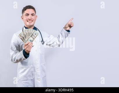 Heureux docteur latin tenant de l'argent et pointant à côté. Beau médecin souriant montrant de l'argent et pointant côté Banque D'Images