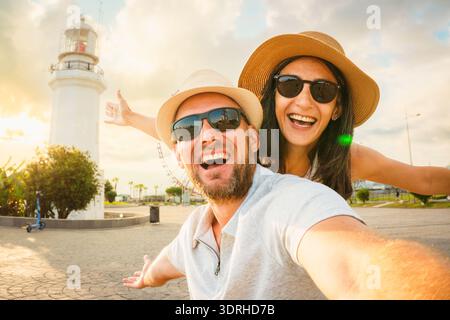 Jeune couple caucasien excité dans l'amour souriant prenant selfie pov à Batumi Boulevard avec phare emblématique et grande roue pendant l'heure d'or. Heureux Banque D'Images