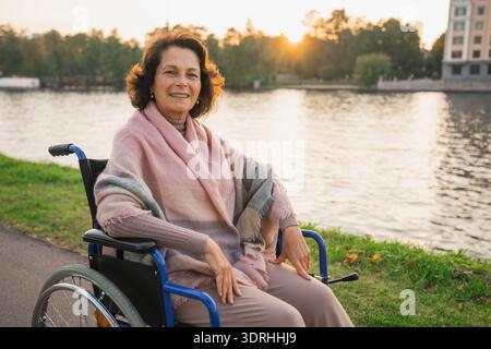 Femme âgée heureuse en fauteuil roulant souriant profitant de la liberté marchant sur la route dans le parc de la ville en plein air. Vieille femme positive joyeuse en c Banque D'Images