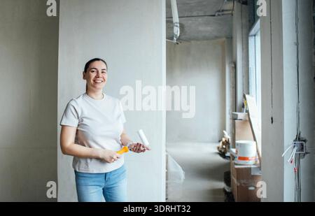 Une femme d'âge moyen tient un rouleau à peinture et sourit. Une jeune dame se tient dans une chambre lumineuse, partiellement rénovée avec w inachevé Banque D'Images