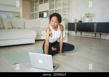 Entraînement de yoga fitness en ligne. Femme africaine pratiquant le yoga avec des leçons en ligne dans un ordinateur portable à la maison. Fille faisant du sport Exe Banque D'Images