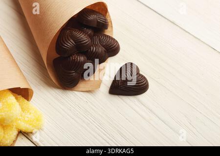 Morceaux de chocolat en forme de coeur dans des cônes de papier avec bonbons gélifiés sur la surface en bois blanc Banque D'Images