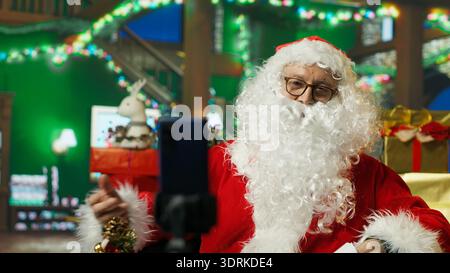 Santa Claus utilisant un smartphone sur trépied pour filmer les lettres d'ouverture de vlog des enfants. Personne âgée portant le costume de Noël enregistrant la vidéo avec la caméra selfie du téléphone, lisant les lettres des enfants, caméra A. Banque D'Images