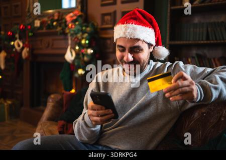 Joyeux Noël. Homme magasinant en ligne tenant smartphone payant avec carte de crédit or pour les cadeaux de Noël. Jeune homme adulte achetant Banque D'Images