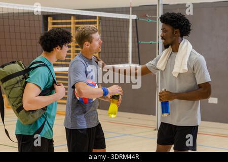 Divers coéquipiers masculins se reposant sur le terrain de volley-ball de gymnastique par filet tenant le volley-ball et des bouteilles d'eau. Athlétique, travail d'équipe, fitness, Banque D'Images