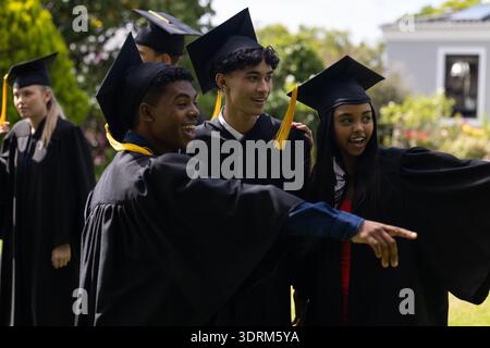 Diplômant les étudiants en casquettes et robes, célébrant à l'extérieur et pointant avec enthousiasme. Obtention du diplôme, éducation, réussite, célébration Banque D'Images