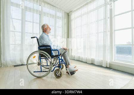 Triste homme âgé assis en fauteuil roulant regardant par la fenêtre dans l'hôpital de la maison de retraite. Vieil homme paralysé dans la chaise pour les gens avec Banque D'Images