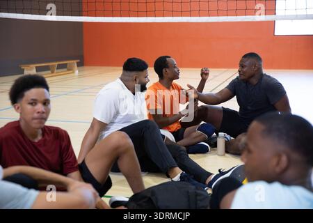 Divers coéquipiers adolescents masculins assis et parlant sur le sol de gymnastique sous le filet avec bouteille Banque D'Images