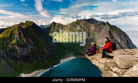 Les aventuriers se détendent sur une corniche rocheuse, en profitant d'une vue imprenable sur la plage de Ryten Kvalvika et les montagnes majestueuses. Verdure luxuriante et dr. Banque D'Images
