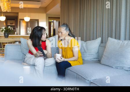 Mère asiatique et fille comparant des photos de smartphone sur canapé gris dans le salon, avec tasse en céramique. Famille, générationnelle, commu Banque D'Images