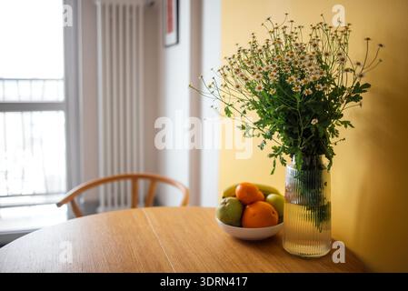 Fleurs fraîches dans un vase clair placé sur une table en bois à côté d'un bol de fruits colorés, créant une cuisine vibrante et accueillante Banque D'Images