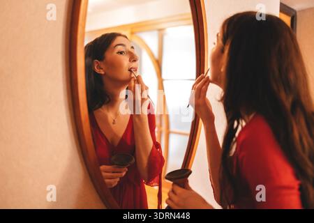 Jeune femme appliquant du rouge à lèvres devant un miroir, vêtue d'une robe rouge, mettant en valeur la routine de beauté et l'auto-soin dans un intérieur confortable Banque D'Images