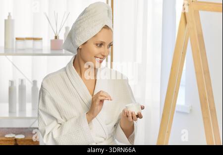 Belle femme portant un peignoir et une serviette enroulée autour de sa tête, regardant la crème petit pot Banque D'Images