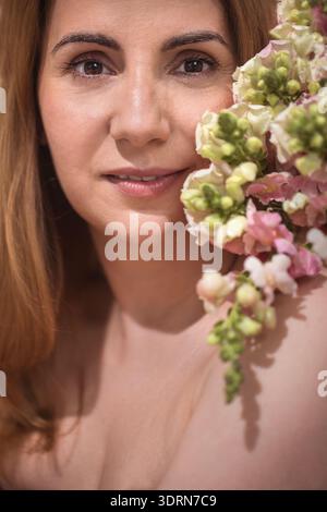 Femme aux longs cheveux bruns, ornée de fleurs colorées, regarde doucement la caméra, incarnant la beauté naturelle et la sérénité Banque D'Images