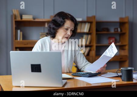 Femme âgée moyenne assise avec un ordinateur portable et un document papier, Pensive vieille dame mature lisant la facture de papier payer en ligne à la maison homme Banque D'Images
