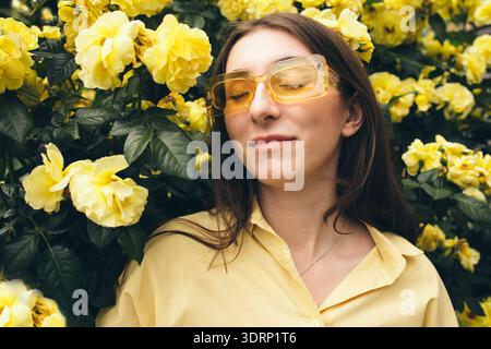 Jeune femme avec de longs cheveux bruns, portant des lunettes de soleil jaunes, pose parmi les fleurs jaunes vibrantes, créant une joyeuse et vivante Banque D'Images