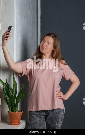 Une jolie jeune femme aux cheveux roux dans un T-shirt clair et un Jean gris se tient contre un mur gris, souriant et posant avec un mobil Banque D'Images