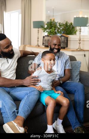 Famille afro-américaine riant tout en touchant les épaules de l'enfant sur le canapé gris dans le salon. Multi-générationnel, bondin familial Banque D'Images