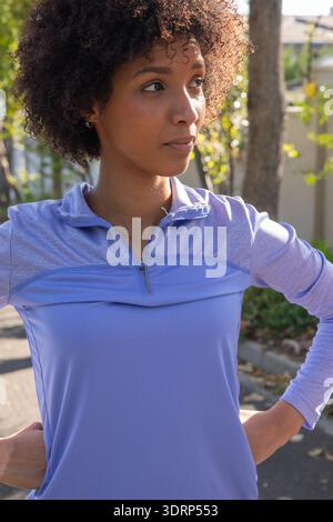 Femme afro-américaine debout sur la rue résidentielle dans le quart-zip de lavande haut, regardant à droite. Vêtements de sport, décontracté, vibrant, ou Banque D'Images