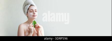 Portrait de beauté de femme en serviette sur la tête avec masque nourrissant blanc ou crème sur le visage et feuille verte à la main, fond blanc i Banque D'Images