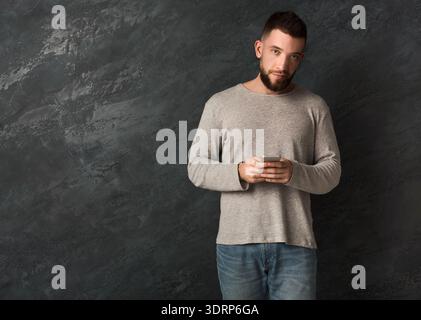 Homme tenant son smartphone tout en se tenant debout contre un mur sombre en intérieur Banque D'Images