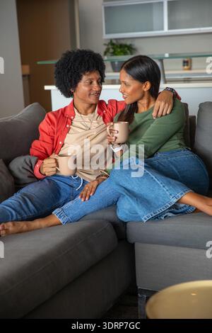 Couple diversifié assis sur un canapé gris dans le salon tenant des tasses en céramique avec plante de cuisine. Intimité, détente, moderne, cosy, Banque D'Images