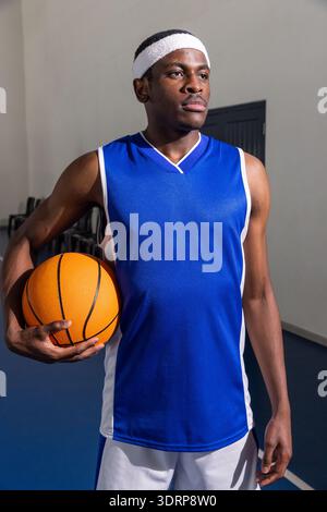 Athlète afro-américain adulte masculin debout sur un terrain de gymnastique portant un maillot bleu tenant un ballon de basket-ball. Athlète, loisirs, actif, s. Banque D'Images