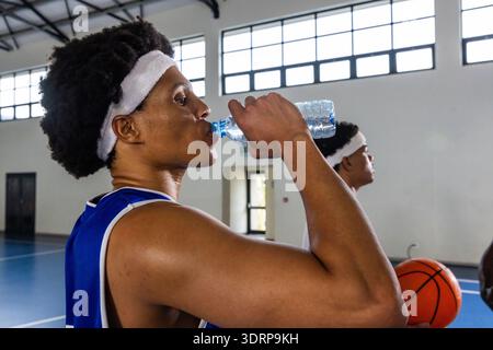 Joueurs afro-américains adultes buvant de l'eau et tenant le basket-ball sur le terrain portant des maillots. Athlètes, fitness, travail d'équipe, Banque D'Images