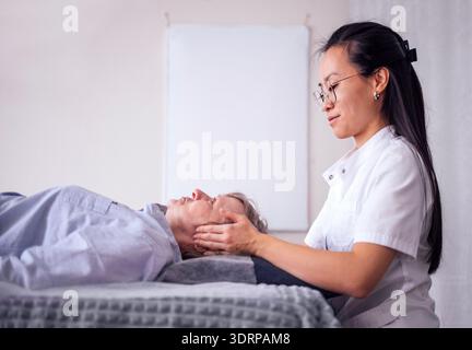 Femme asiatique offrant une massothérapie faciale à un client senior dans un studio de bien-être serein, mettant en valeur la relaxation et le rajeunissement Banque D'Images