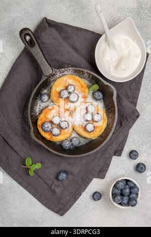 Crêpes au fromage cottage, syrniki, beignets de lait caillé avec baies fraîches, myrtille, sucre en poudre et crème sure dans une poêle en fonte. Banque D'Images