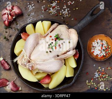 Le poulet cru de ferme entier avec pommes de terre, ail, thym et épices (poivrons et gros sel) est prêt à cuire dans une poêle en fonte. Banque D'Images