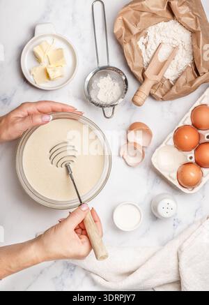 Femme prépare la pâte pour les crêpes maison pour le petit déjeuner. Fouet pour fouetter dans les mains. Ingrédients sur la table - farine de blé, e Banque D'Images