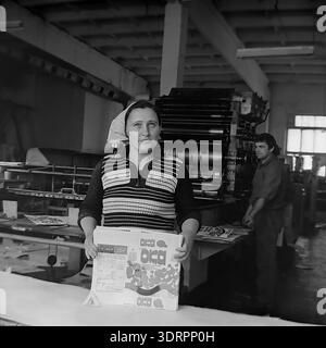 Une photographie historique en noir et blanc des années 1960 capture une ouvrière d'usine tenant une pile d'emballages en carton fraîchement imprimés pour la poudre à laver Oka à l'usine d'huile et de graisse de Slavyansk en RSS d'Ukraine, URSS. Vêtue d'un pull rayé et d'un foulard, elle se tient debout dans l'atelier d'imprimerie tandis qu'un collègue masculin exploite une grande presse offset dans le fond flou. Cette image d'archives authentique met en évidence l'ampleur de l'autosuffisance industrielle soviétique, la dignité du travail et l'atmosphère optimiste dans le Donbass pacifique avant la guerre. Banque D'Images