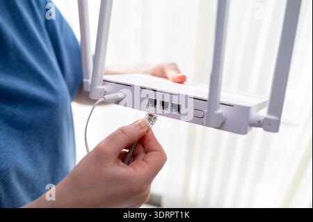 Gros plan d'une personne en chemise bleue connectant un câble réseau à un routeur sans fil. Ce cliché représente l'installation internet à domicile, le tro technique Banque D'Images