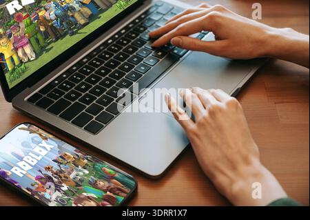 Riga, Lettonie - 2 février 2026 : gros plan des mains à l'aide d'un ordinateur portable alors que Roblox est affiché à l'écran, avec un smartphone montrant le même jeu sur un WO Banque D'Images