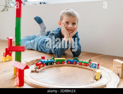 Enfant souriant allongé sur le sol et jouant avec le train jouet en bois mis à la maison. Ambiance familiale confortable, développement de la petite enfance et intérieur créatif Banque D'Images