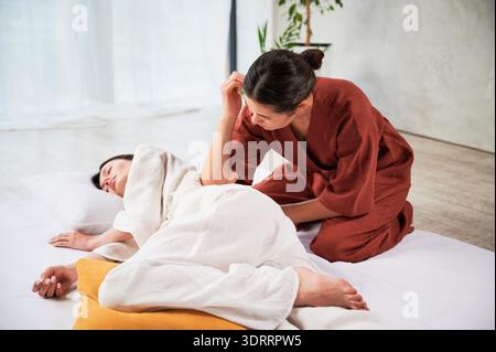 Masseuse faisant massage yoga thaïlandais. Thérapeute de femme faisant le traitement de massage traditionnel pour la patiente féminine utilisant le coude. Client allongé sur tapis, méditant, profitant d'un massage des mains dans une atmosphère calme. Banque D'Images