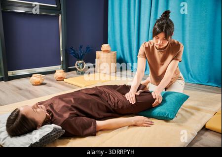Masseuse faisant massage yoga thaïlandais. Thérapeute de femme faisant le traitement de massage traditionnel, étirant le corps de la patiente féminine. Client allongé sur tapis, profitant d'un massage des mains dans une atmosphère calme. Banque D'Images