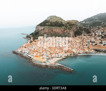 Vue aérienne des toits en terre cuite en cascade vers la mer Azur, nichée sous la majestueuse Rocca di Cefalù, créant un contraste de couleurs époustouflant Banque D'Images