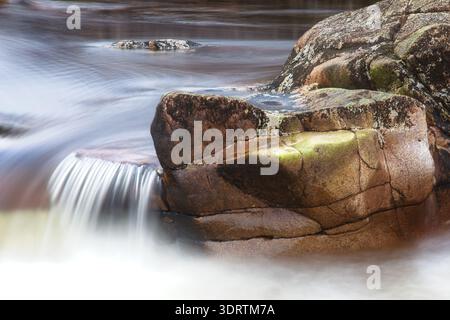 Vitesse lente de la navette d'une petite cascade coulant sur un rebord de roche moussue et des rochers dans une rivière écossaise. Banque D'Images