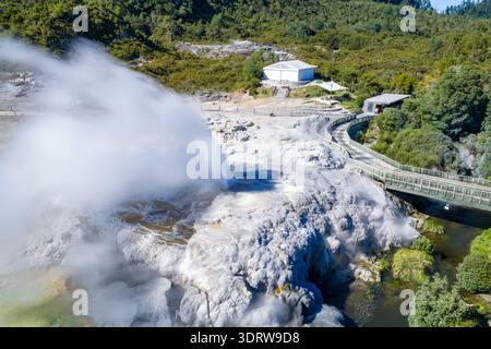 Vue aérienne d'un geyser en éruption puissante, envoyant un panache de vapeur blanche dans l'air près d'un pont et d'un bâtiment, Rotorua, région de la baie de Plenty, ne Banque D'Images