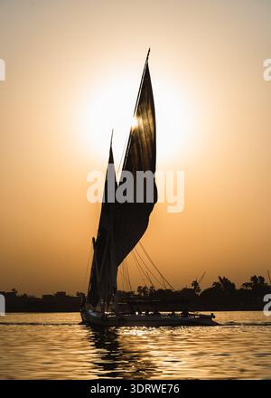 Vue d'un bateau felouque naviguant sur les eaux tranquilles et réfléchissantes du Nil, ses voiles silhouettées contre le soleil couchant, Louxor, gouvernorat de Louxor Banque D'Images