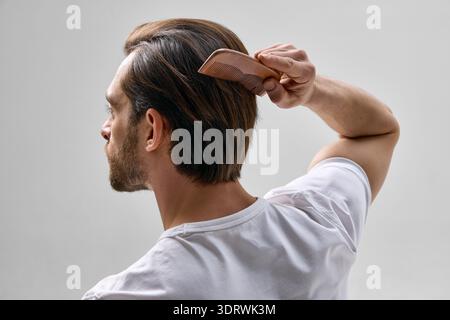 Vue arrière de l'homme dans la trentaine avec la barbe courte brossant les cheveux avec peigne en bois sur fond clair, portant t-shirt blanc dans un cadre minimal. Banque D'Images