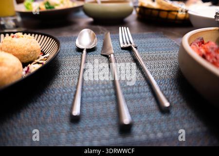 Fourchette de couteau cuillère en acier inoxydable disposée sur une nappe texturée bleue avec des assiettes de nourriture en arrière-plan Banque D'Images