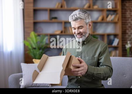 Homme senior avec les cheveux gris et la barbe assis sur un canapé, ouvrant un colis de carton à la maison, éprouvant la joie et la satisfaction de son nouvel achat ou cadeau d'une commande en ligne Banque D'Images