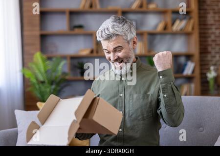 Homme mûr avec une barbe grise et une chemise assis sur un canapé, déballant avec joie un paquet de carton, montrant l'excitation et le succès avec une pompe à poing après avoir reçu sa commande anticipée Banque D'Images
