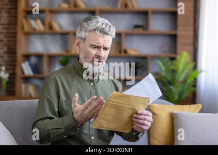 Homme mûr aux cheveux gris assis sur un canapé, tenant une enveloppe ouverte avec une expression inquiète et frustrée après avoir lu un courrier financier ou officiel pénible à la maison Banque D'Images