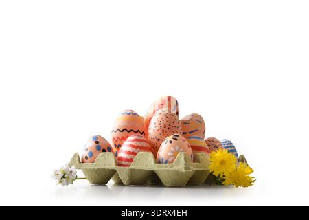 Un carton rempli d'œufs de Pâques peints à la main décorés de motifs lumineux et de points, placé sur une surface blanche avec des pissenlits jaunes et de petits blancs Banque D'Images