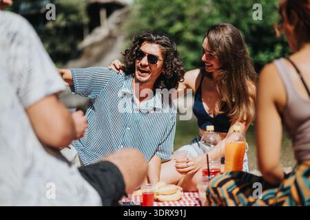 Amis profitant d'un pique-nique ensoleillé au bord de l'eau, souriant et riant ensemble Banque D'Images